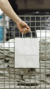 persoon houdt witte papieren draagtas met koordhandvat boven stapel draagtassen in magazijn